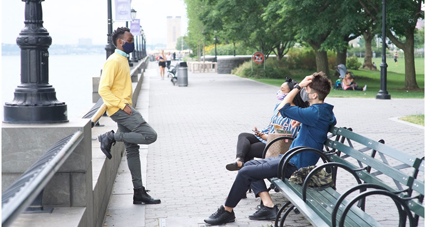 three students, two on a bench and one standing, talking, along a path with trees near campus