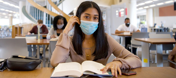 female student wearing mask sitting at desk with open textbook