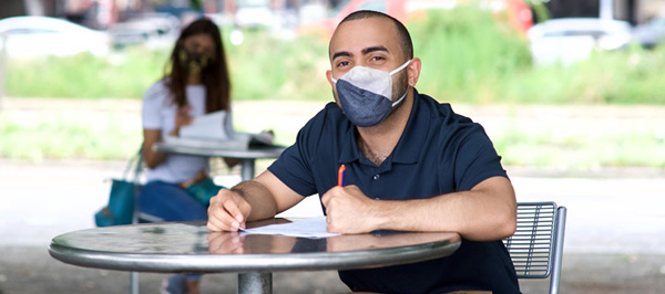 student wearing mask outside at picnic table with pen and papers