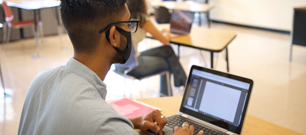 student wearing mask using computer in a classroom with other students, socially distanced