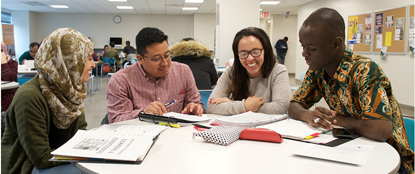 four students sitting at table looking at assignments and talking